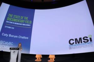 Caty Borum Chattoo, a white woman standing behind a podium, presenting the 2018 State of the Documentary Field Report. A presentation screen behind her. Courtesy of CMSI