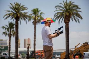 John Wilson is a white male filmmaker wearing a colorful sun umbrella around his head. He is holding a video camera with palm trees in the background. From ‘How To with John Wilson.’ Image courtesy of Joe Buglewicz/HBO.