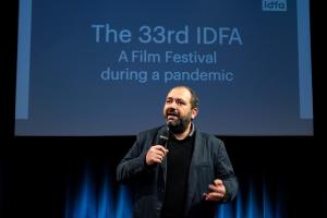 The IDFA festival director, mic in hand and wearing a black jacket, addresses the audience in front of a blue curtain.