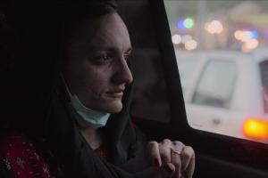 Zarifa Ghafari, an Afghan woman with black shoulder-length hair, wearing a black coat and a red patterned scarf and a surgical mask around her neck, is sitting in the back seat of a moving car, looking out the window. From Tamana Ayazi and Marcel Mettelsiefen’s In Her Hands. Courtesy of Netflix ©2022