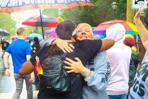  A mother from the mama bears movement hugs a person wearing a black backpack with rainbow pride flags hanging from it during a pride celebration. Photo courtesy of Mama Bears.