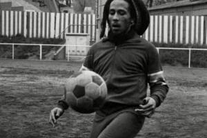 Reggae legend and football fan Bob Marley, in dreadlocks and beard and wearing a dark jersey, dribbles a football. From ‘Legacy:Rhythm of the Game.’