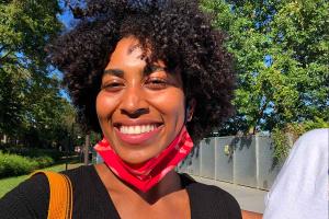 A Black woman with a huge smile and short curly hair is seen wearing a red mask on half of her face in a green and luscious park.
