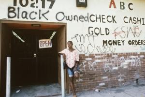 A Black woman leans against a wall that is spray painted with phrases like, 'Black owned." From 'No Loans Today.