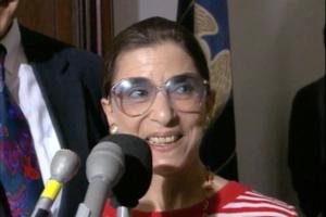 Justice Ginsburg is pictured here at a press conference, in front of a bank of microphones, and she is wearing a red-and-white striped shirt.