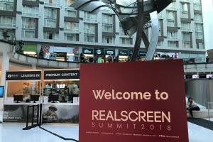 A red sign with "Welcome to REALSCREEN SUMMIT 2018" in front of an open-air business lobby with abstract metal sculpture.