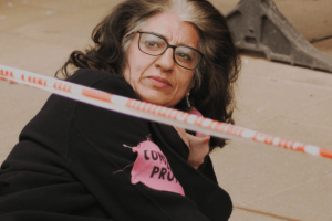 A woman protestor, with shoulder-length brown hair, dressed in black and wearing glasses, is sitting on the ground looking over her shoulder. From Maia Kenworthy & Elena Sanchez Bellot's “Rebellion.” Photo courtesy of DOK Leipzig. 