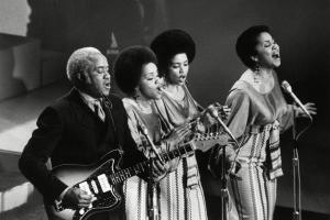 Three Black women in long skirts singing, one Black man playing the guitar. They are on stage. There are three microphones.