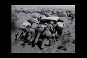 A black-and-white photo of a group of people pushing a car through a barren landscape. 
