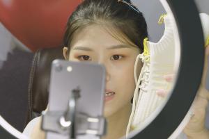 An asian woman with black hair holds up a white tennis shoe in front of a camera and ring light.