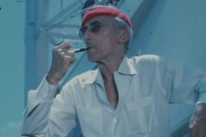 An older white man wearing a red cap, white shirt and sunglasses. He is smoking a pipe.