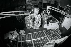 A black and white photo of an asian man in a patterned button up. He sits in a recording studio.