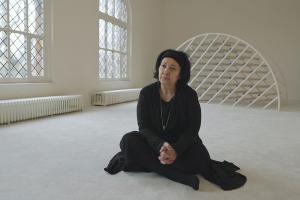 An older woman with brown skin and black hair wearing a black outfit sits on the floor.