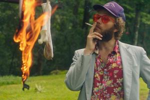 A white man with brown hair and facial hair wearing red shades and a Hawaiian shirt and blazer. He smokes a cigarette and watches socks burn.