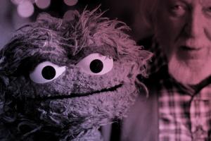 An older white man with gray hair holds up a green, scraggly puppet.