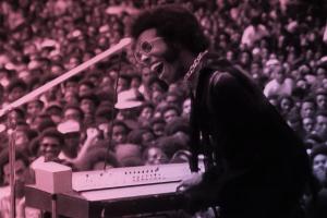 A Black man with black hair wearing pink glasses, a gold necklace and a purple suit. He is playing keyboard to a crowd.