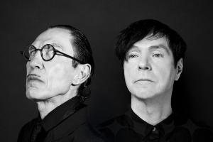 A black and white portrait of two white men both with dark hair. The man on the left has round black glasses and a pencil mustache.