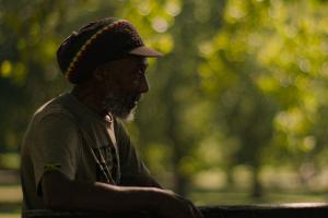 Reggae musician sits before a lush green forest. 