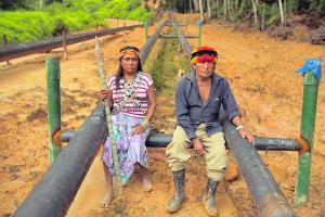 Two indigenous people sitting on black pipes in the Amazonian forest.