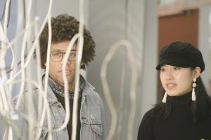 A young Black man with curly hair, glasses and a scarf and a young Asian woman with a black cap and white earrings stand before an abstract white curly art piece in a museum.