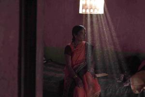 An Indian woman in a sari sits on a couch before a window. Light rays stream through the window's elaborate design