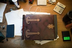 A rustic old notebook of archived documents sits on a desk surrounded by smaller papers and notepads.