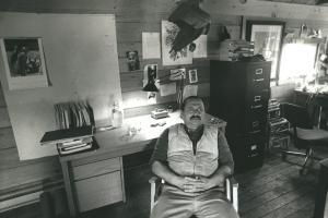 Jim Harrison sits with his hands in his lap in his rugged, outdoorsy 20th-century office. 