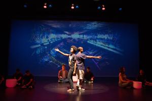 Two young Black teenagers stand back-to-back with a single arm stretched out in opposite directions. They stand under a spotlight on stage in front of a projection screen showing a graffitied pipeline. Several other students sit on the stage around them, sitting next to sound bowls.