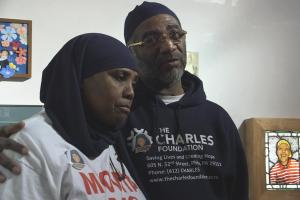 A still from "Murders That Matter": Protagonist Movita Johnson-Harrell, a middle-aged Black woman in a white t-shirt and black hijab, stands next to a middle aged Black man wearing a blue hoodie that says "The Charles Foundation"