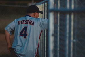 Baseball player Lenny Dykstra leans against a wire fence. He is wearing a white jersey that says "Dykstra - 4"