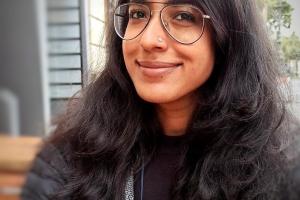 Woman of Indian descent with long black hair and glasses takes a selfie with a blurred background