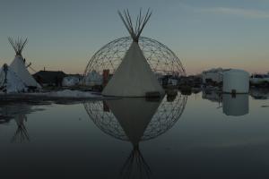 a white tipi sits in front of a steel geometric dome, both are reflected in a shallow pool at sunset.