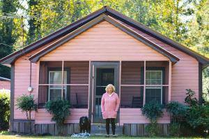 An older woman in a pink cardigan stands in front of a pink house with her dog.