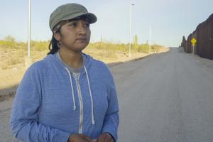 An indigenous woman stands in the shadow of the US-Mexico Border Wall