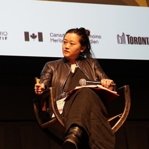 A woman with black hair sits at the chair of a panel and is mid-speech. 