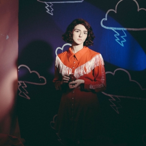 A woman with short black hair stands in front of a dark blue backdrop with clouds on it. She wears a red cowgirl outfit. 