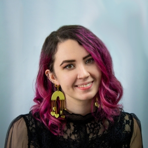 A white woman tilts her head and smiles at the camera against a light blue backdrop. Her hair is purple and she wears large earrings. She was an elegant black lacy top.