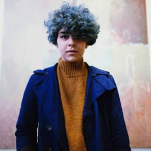 A person with blue curly hair looks straight into the camera. They wear a mustard colored turtle neck and a bright blue blazer. 