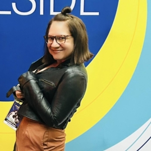 A woman stands in front of a colorful back drop at a film festival. She has shoulder length brown hair and glasses. She wears a tight black leather jacket and an orange skirt. 