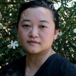 Portrait of Abby Sun, IDA's director of programs and editor of Documentary magazine. An Asian woman with short hair, wearing a black blouse and standing in front of a white flower bush.