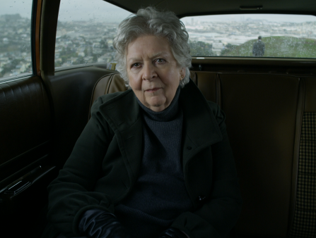 In a car interior, a white woman with short-cropped silver hair looks archly at the camera.