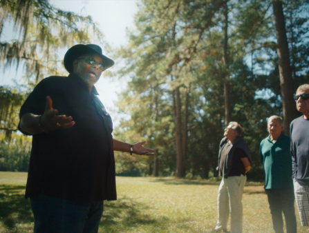 A Black man talking to a group of white people, while standing in the middle of a forested area.