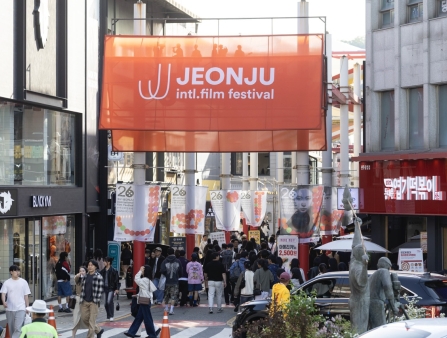 On a busy street, the orange banner for the Jeonju International Film Festival is pictured above pedestrians and cars.