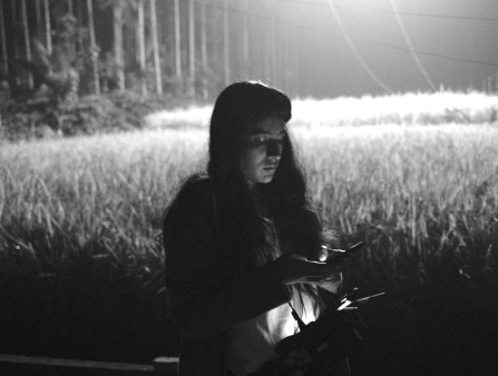 Dark, black and white photo of a woman with long hair looking into a monitor screen at night, under a bright floodlight in a field.