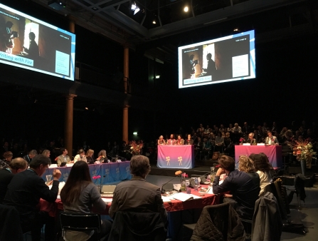 In a large room surrounded by tables, four people sit a small table below two large screens.