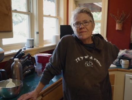 An older woman in a hoodie leans against a kitchen counter.