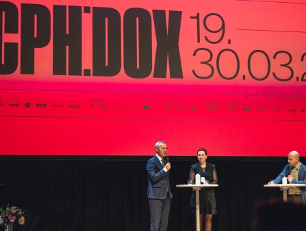 In front of a red screen, three people stand at cocktail tables, holding microphones.