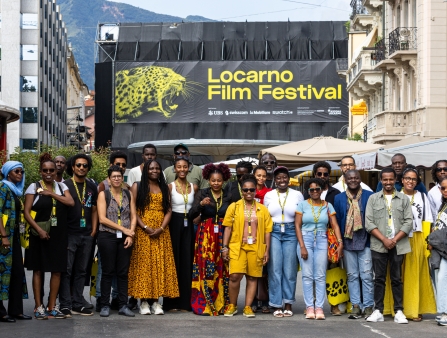 A large group of smiling people wearing Locarno merchandise pose in front of a festival banner.