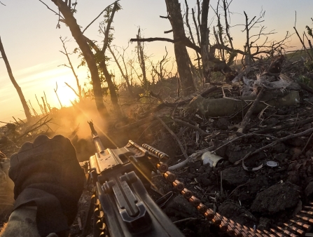 From the point of view of a soldier's helmet, a machine gun is in focus as the sun comes up over a destroyed field