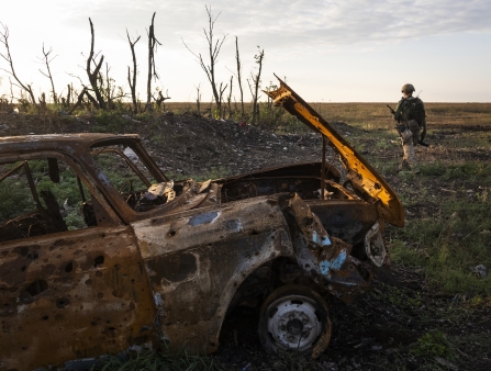 A burnt car is seen in foreground while a soldier nearby is seen in the distance in an empty field
